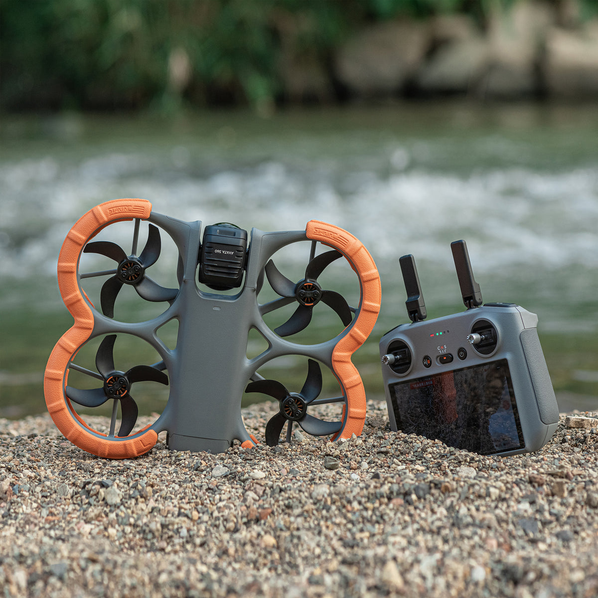 DJI Avata 360 equipped with STARTRC EVA bumper guards resting on gravel near water, alongside the remote controller. Essential DJI Avata 360 accessories for outdoor protection.